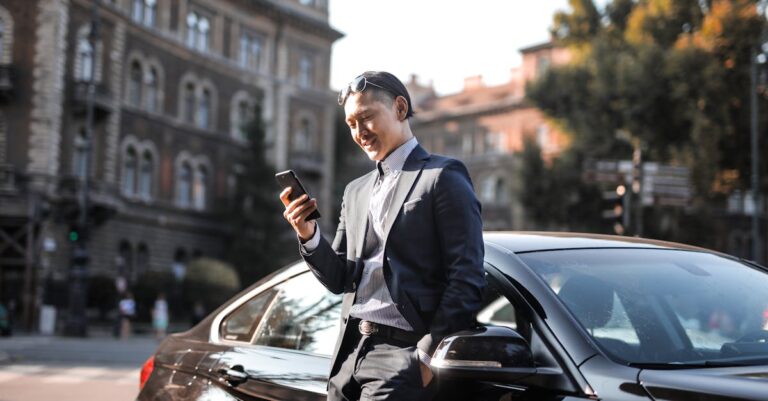 Selective Focus Photo of Man in Black Suit Using His Phone While Leaning Beside Black Car