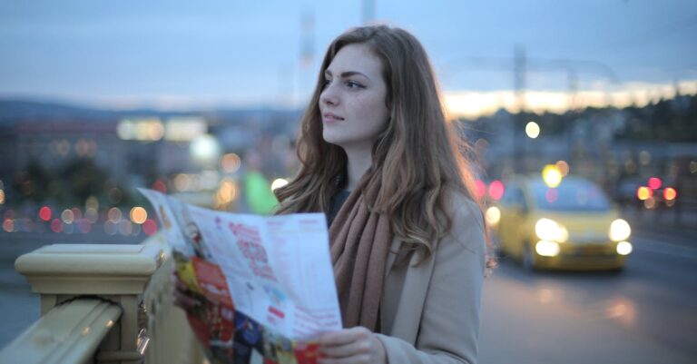 Female traveler in warm clothes standing on street with map and looking away at dusk