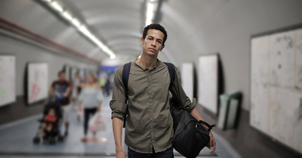 Calm young African American male in casual clothes with big black bag and backpack looking at camera while walking along corridor of underground station against blurred passengers