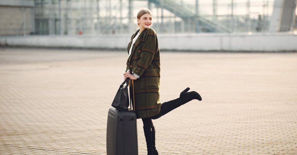 Full body of stylish female tourist in warm clothes and high heeled boots standing on background of blurred airport terminal with luggage and ladies bag