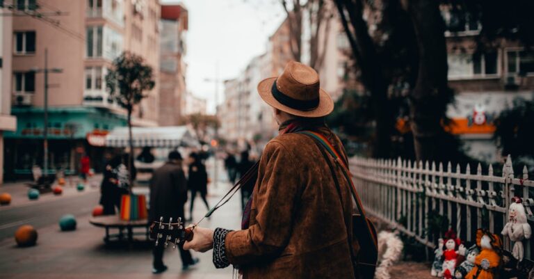 Man in Brown Jacket and Brown Hat Standing on Sidewalk With A Guitar