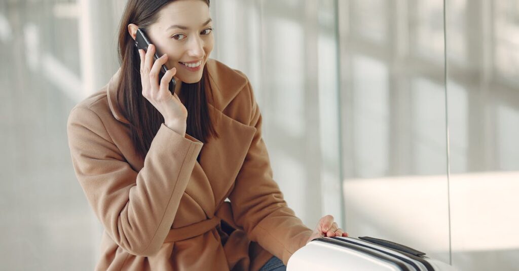 Young dreamy brunette in beige coat with suitcase having conversation via mobile phone while waiting for flight in airport terminal