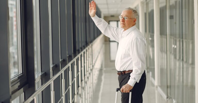 Side view of aged calm male in formal clothes with suitcase walking along modern airport hallway and waving goodbye while looking out glass wall