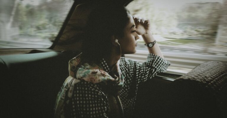 Side view of pondering young ethnic lady listening to music in earphones while sitting leaned on hand near window inside automobile and looking away