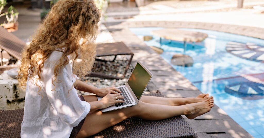 From above side view of unrecognizable barefoot female traveler with curly hair typing on netbook while resting on sunbed near swimming pool on sunny day