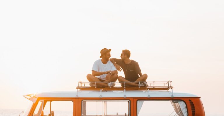 Silhouettes of full body faceless male friends in casual clothes resting on camper van roof and chatting in countryside in sunset time