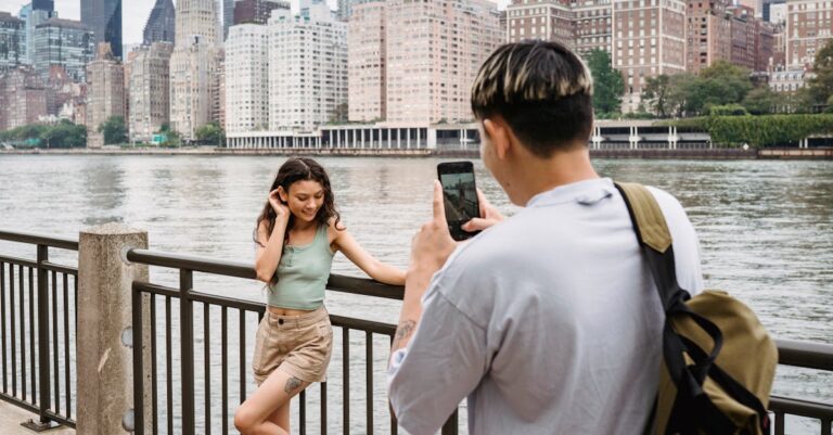 Young man photographing girlfriend on smartphone during date in city downtown near river