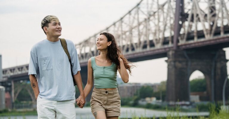 Happy ethnic couple holding hands while strolling in park near bridge