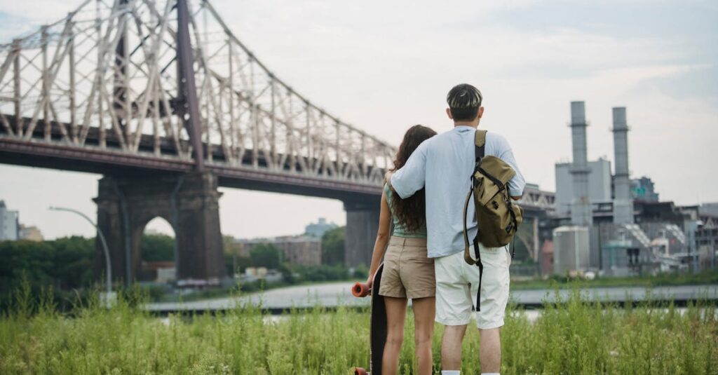 Back view of unrecognizable young male tourist in casual clothes with backpack cuddling girlfriend with skateboard in hand while standing on grassy ground near Brooklyn Bridge and admiring city