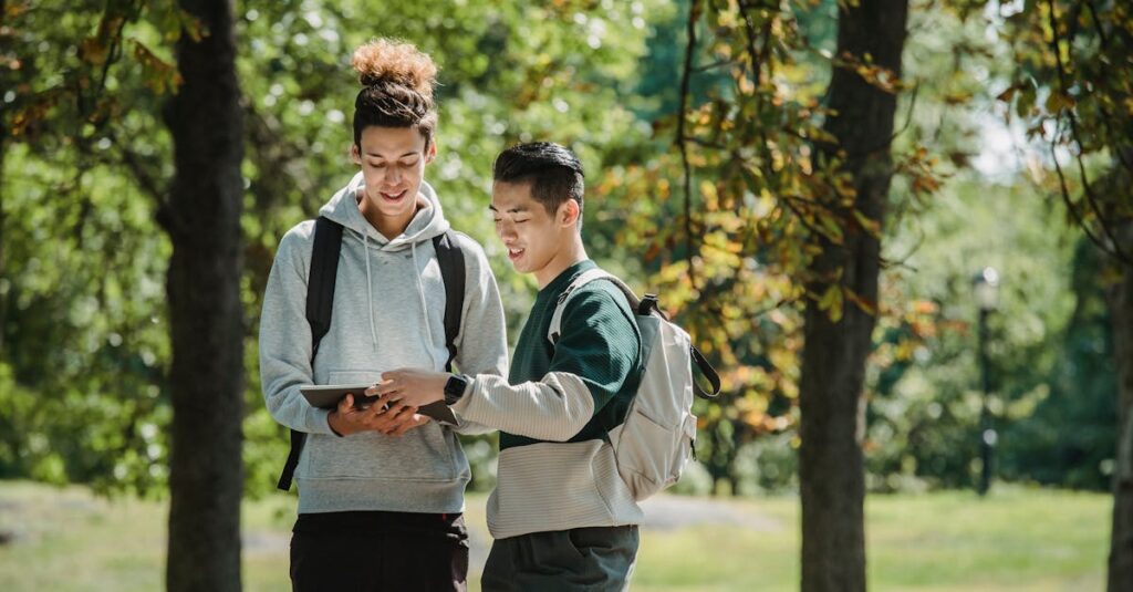 Confident young multiracial male friends in trendy outfits and backpacks standing in green park on sunny day and reading notebook together before studies