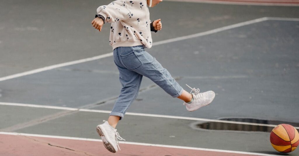 Side view of jumping little girl in jeans and jumper kicking colorful ball playing on sports yard