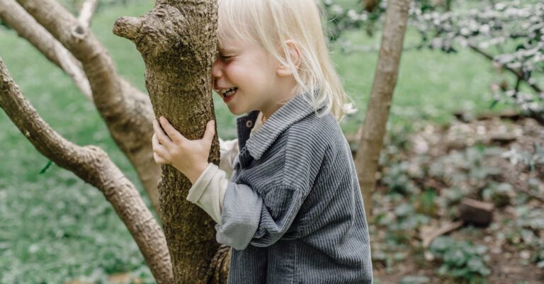 Side view of cheerful little kid with closed eyes smiling and touching tree trunk while standing in park on blurred background