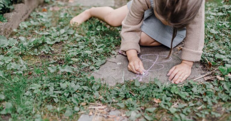 High angle of crop unrecognizable barefooted child painting on ground with colorful chalks while playing in green garden