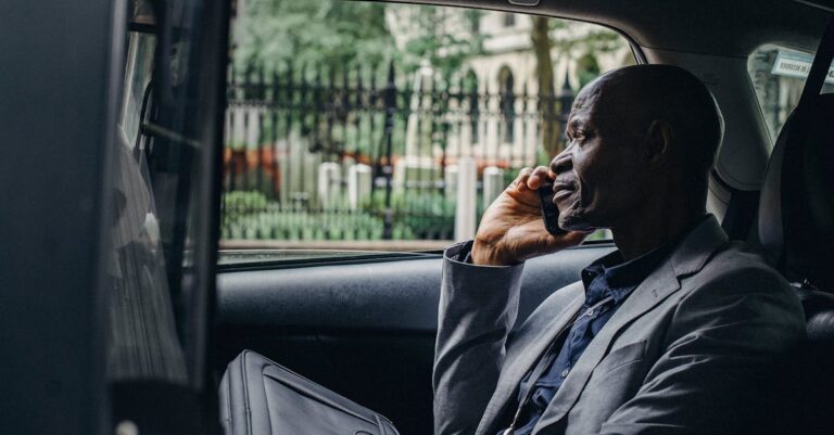 Side view of thoughtful mature African American businessman in elegant suit talking on smartphone while riding on passenger seat of taxi