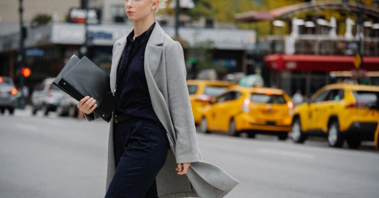 Young confident well dressed blond female entrepreneur with folders walking on asphalt roadway while looking forward in town