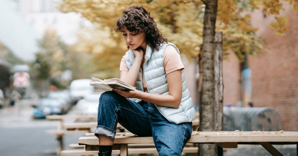 Pensive female in trendy outfit reading book while sitting on wooden table near autumn tree on city street in daytime