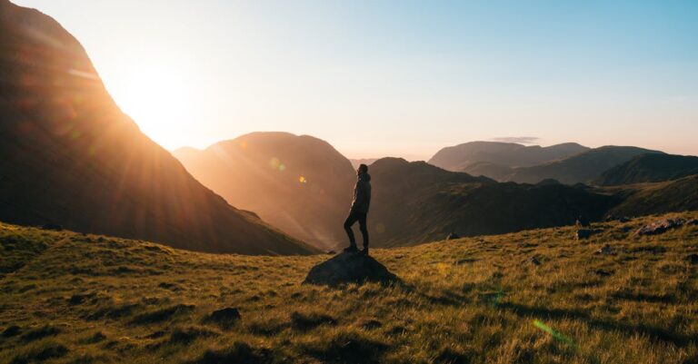 Silhouette Photography of Person Standing on Green Grass in Front of Mountains during Golden Hour