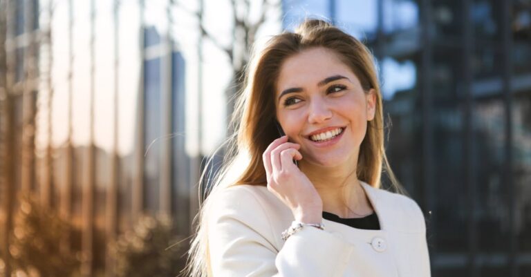 Selective Focus Photo of Woman in White Long Sleeve Coat Smiling While Talking on the Phone