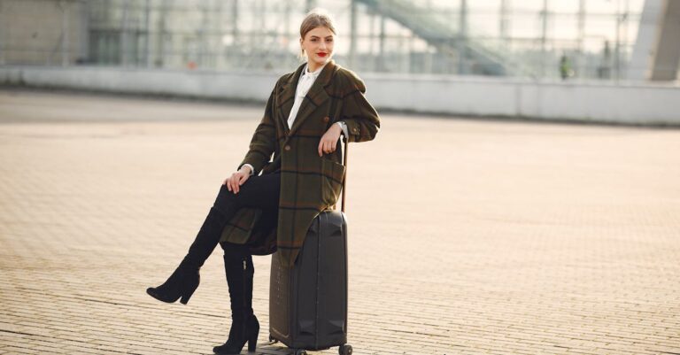 Full length of positive female traveler wearing warm trendy plaid coat and high boots sitting on suitcase with legs crossed on pavement near modern airport terminal and smiling looking at camera in daylight during cold season