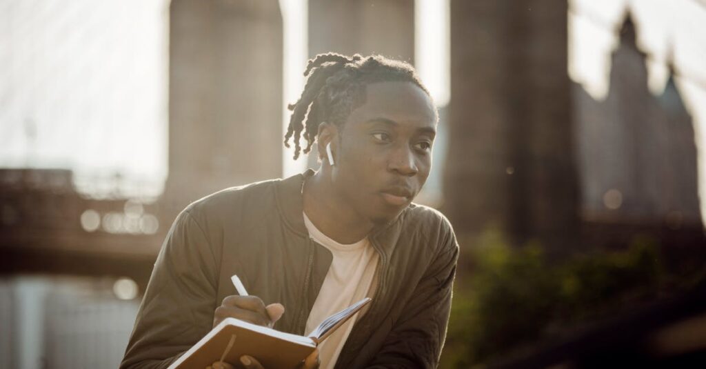 Pensive black man drawing picture in notebook while standing on riverbank