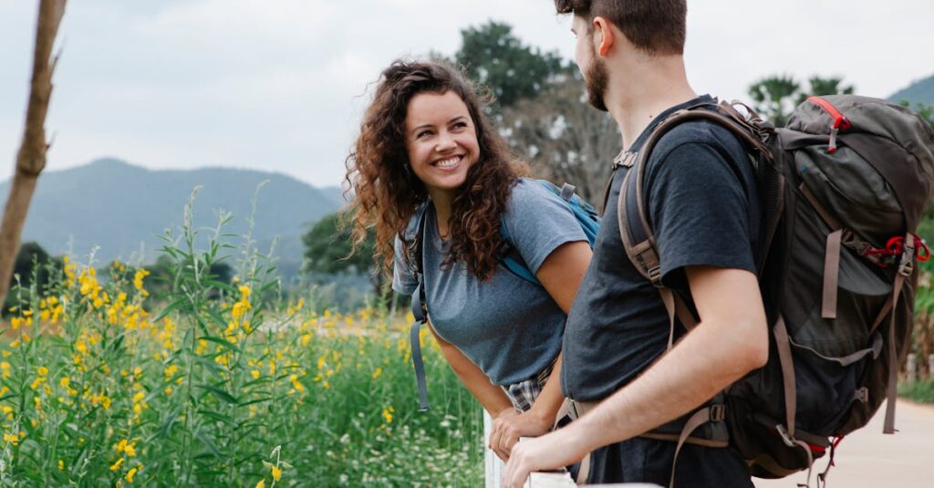 Happy young couple of tourists in casual clothes with backpacks looking at each other while leaning on wooden fence near grassy field with wildflowers in daytime near mountains and trees