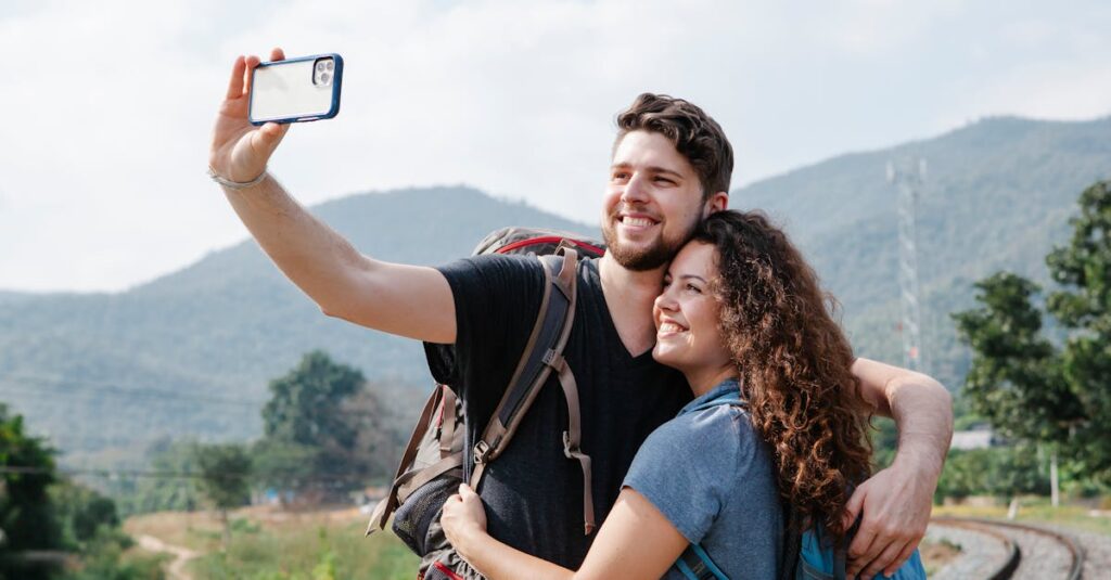 Cheerful couple of tourists in casual clothes with hiking backpacks hugging and taking selfie on smartphone in nature against green hills in daytime