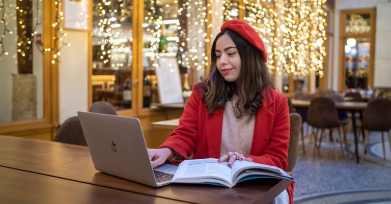 Young woman in red beret studying with laptop in a Parisian café with festive lights.