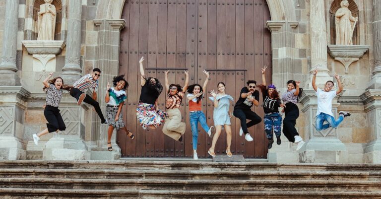 A diverse group of young friends joyfully jumping on stairs in front of a historic building entrance.