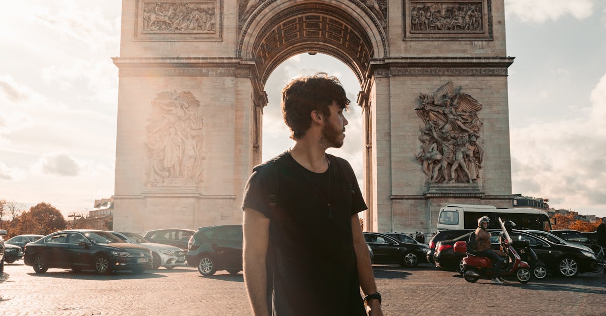 A stylish man poses in front of the iconic Arc de Triomphe in Paris, showcasing fashion and architecture.