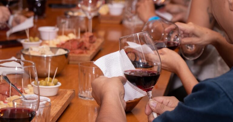 Close-up of people's hands holding wine glasses at a wine tasting event with snacks on a wooden table.