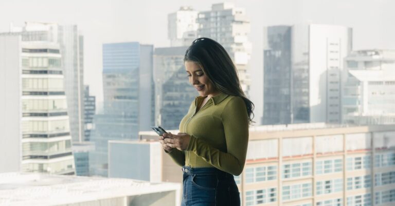 Woman looks at smartphone inside high-rise building with cityscape in background.