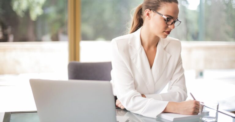 Focused businesswoman in white suit taking notes at a desk in an elegant office setting.