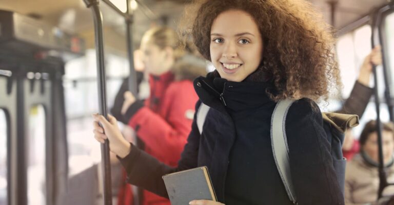 A young woman stands with a book, smiling on public transport during daytime travel.