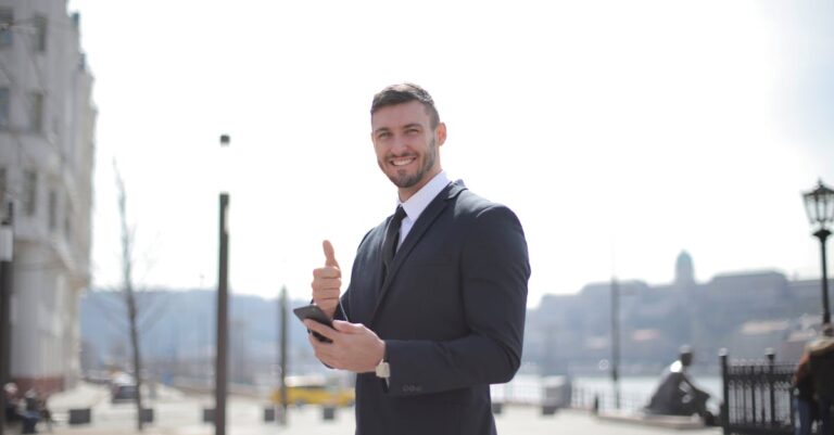 Smiling businessman in a suit giving thumbs up and holding a smartphone outdoors in a city setting.