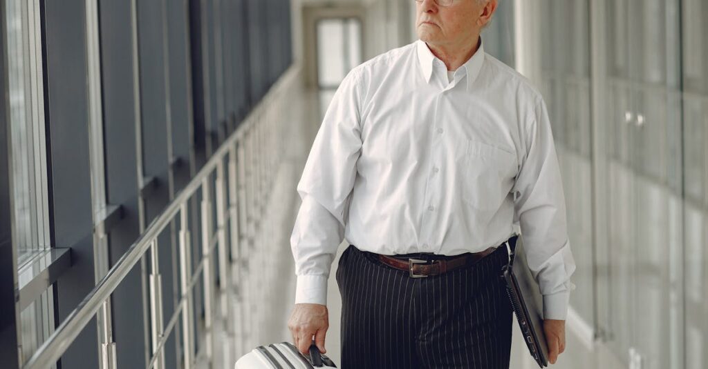 Serious senior male in formal clothes walking along airport corridor with baggage and laptop while looking away through window