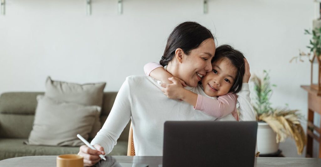 Happy young Asian woman working remotely from home with laptop and tablet while adorable little daughter hugging from behind