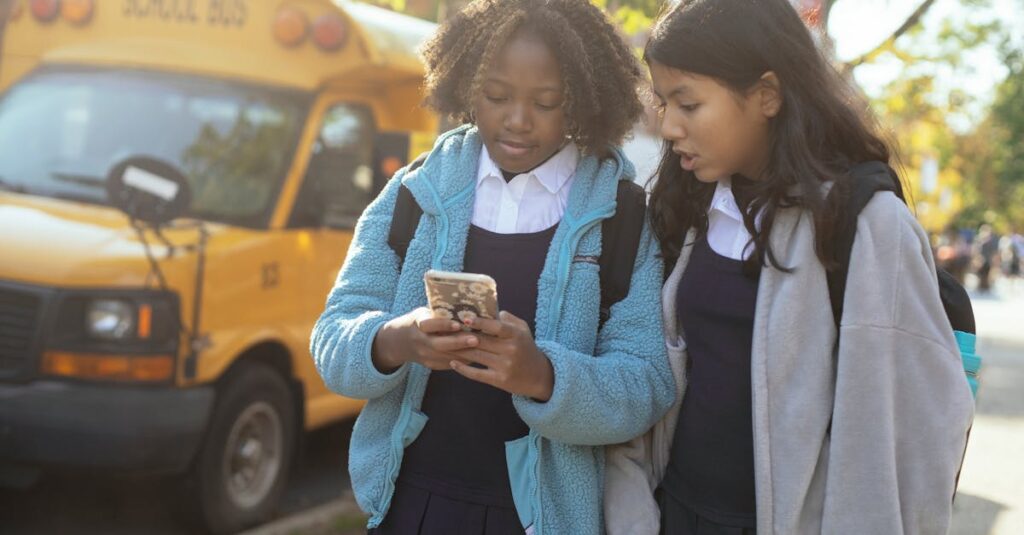 Two schoolgirls using a phone near a school bus, enjoying a sunny day, showcasing friendship and technology.