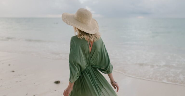 Back view of unrecognizable female tourist wearing straw hat and summer dress standing on seashore in cloudy day