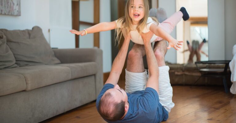 Joyful dad bonding with daughter in playful airplane pose indoors.