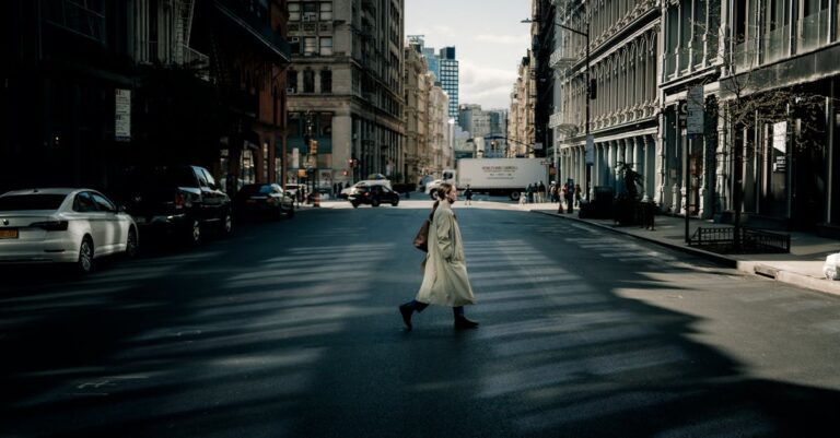 A woman in a trench coat walks across a sunlit street in New York City with tall buildings surrounding her.