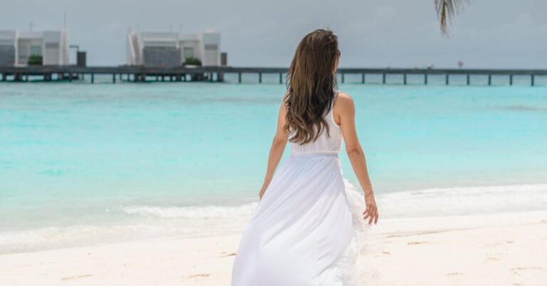 A woman in a flowing white dress walks on a pristine tropical beach with turquoise waters.