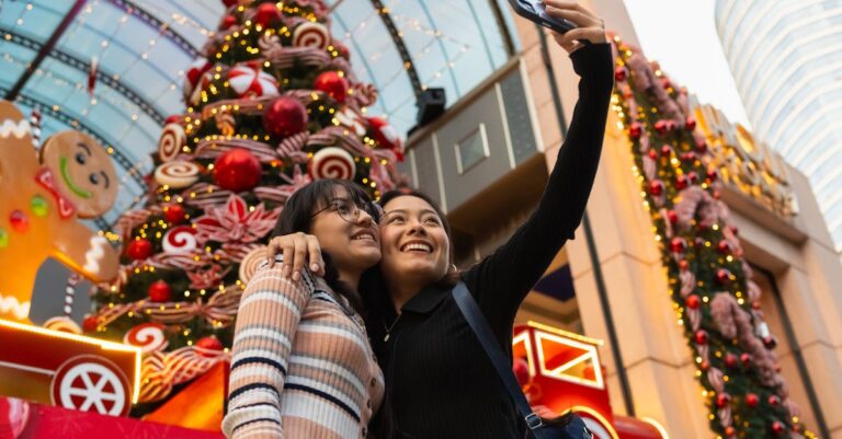 Two women taking a selfie in front of festive Christmas decorations at a mall.