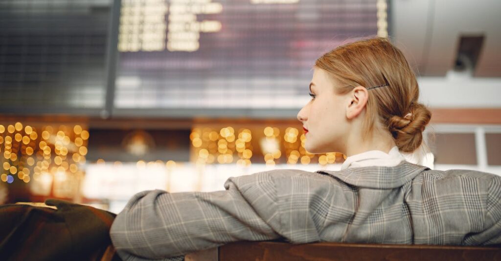 Pensive female designer in checkered jacket waiting for flight near departure board looking away in modern airport in evening