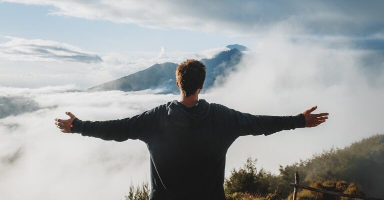 Back view of anonymous male traveler in casual clothes standing on edge of cliff and admiring breathtaking scenery of clouds and mountain top