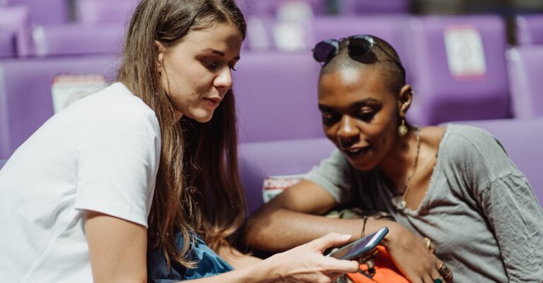 Two adult women sitting together and sharing a smartphone in a public waiting area with purple seats.