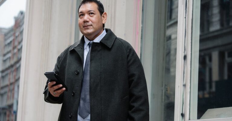 Low angle of confident adult ethnic male manager in classy suit and coat standing on street near building and browsing mobile phone