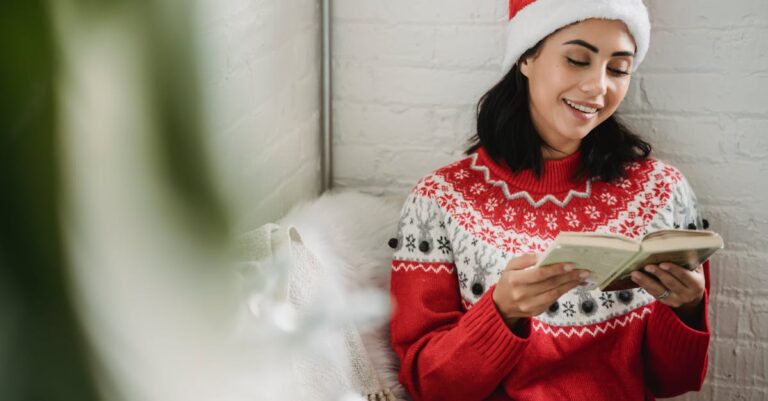 Woman in festive sweater and Santa hat enjoying a book indoors during winter holidays.