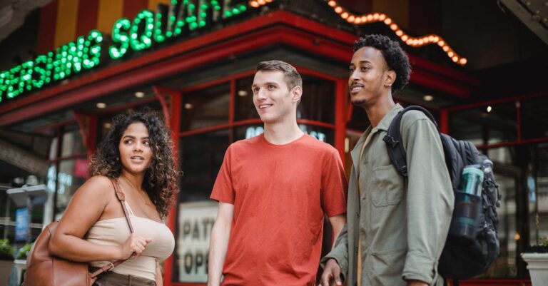 Group of young multiracial friends in casual outfits with backpacks standing in city street near building with sign and looking away in daytime