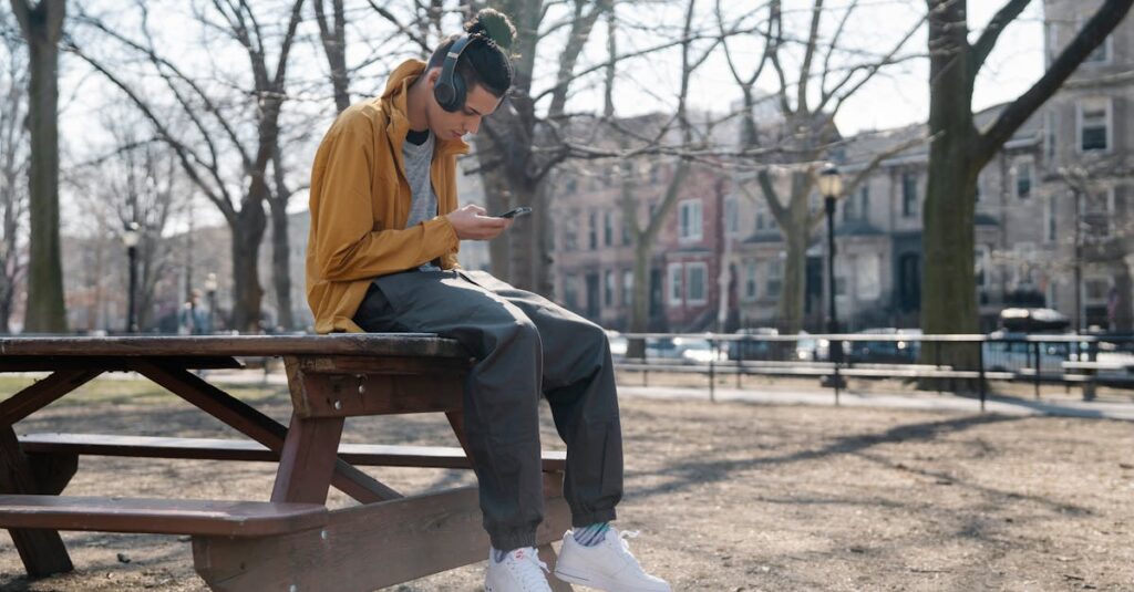 Cool ethnic male in headphones text messaging on cellphone while listening to music and sitting on table in town