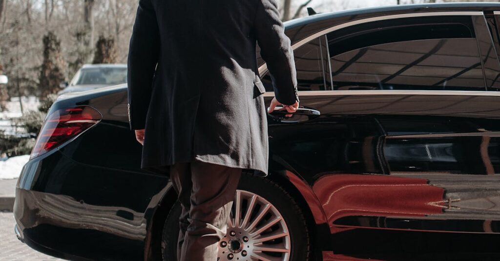 Bald man in a black suit approaches a luxury black car on a sunny day.